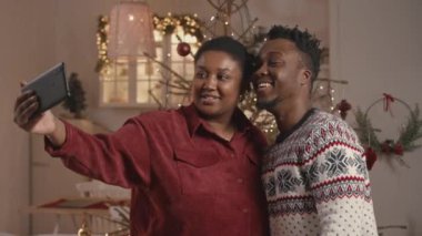 Orta karar Afro-Amerikan çiftin Noel için hazırlanmış rahat evlerinde süslü Noel ağacıyla akıllı telefonlarında selfie çekerken orta boy bir fotoğrafı.
