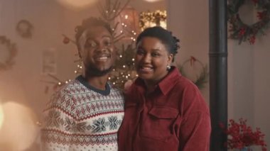 Orta mercekli genç Afro-Amerikan çiftin fotoğraf makinesi için poz veren portresi Noel için güzel süslenmiş sıcak ve rahat bir dairede çelenkler ve çelenklerle dikildi.