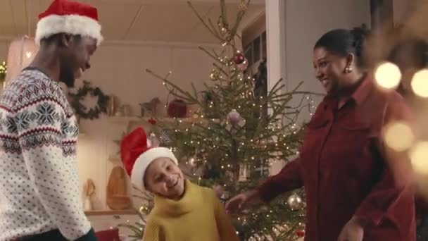 赤いサンタハットダンスで喜びのアフリカ系アメリカ人の女の子のミディアムショットとクリスマスイブの自宅で両親と楽しんでしながらウィリング — ストック動画