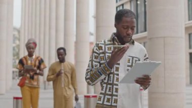 Genç Afrikalı Amerikalı bir adamın iş belgeleriyle dolu klasöre bakarak sokakta yürüyüp akıllı telefondan sesli mesaj kaydettiği orta ölçekli bir görüntü.