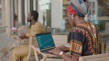 Geleneksel kostüm ve türbanla dizüstü bilgisayarda çalışan, gündüzleri açık kafede oturan Afro-Amerikan genç kadının arka görüntüsü.