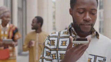 Geleneksel giysiler içinde genç Afrikalı Amerikalı iş adamının şehir merkezindeki caddede yürürken akıllı telefondan hoparlörden konuşmasını göğüsleyelim.