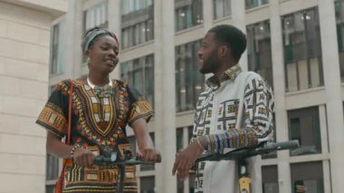 Düşük açılı orta açı çekimde geleneksel canlı giysiler içinde genç Afrikalı Amerikalı bir çift sokakta motosikletlerinin yanında durup dostça sohbet ediyorlar.