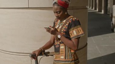 Arican 'lı genç bir Amerikalı kadının güneşli bir günde sokakta dikilirken ve akıllı telefondan bakarken orta boy fotoğrafı.