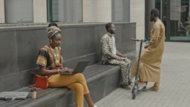 Sarıklı ve geleneksel giyimli çekici genç siyahi kadın yaz günü şehir merkezinde bankta otururken dizüstü bilgisayarda çalışıyor.