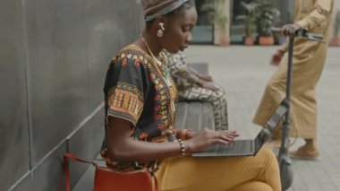 Sarıklı ve çağdaş geleneksel giysileri olan genç Afrikalı Amerikalı kadın şehir ortamında dizüstü bilgisayarla çalışıyor.