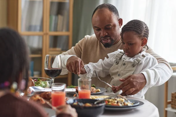 African dad sitting at table with child on his knees and helping her to eat meal during family dinner