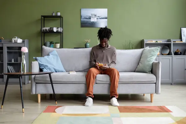 African young guy sitting on sofa in living room and communicating online using his mobile phone