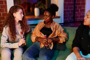 Kültürler arası üç genç kadın ellerinde bira şişeleriyle oturma odasındaki kanepede dinleniyorlar ve ev partisinde dans ettikten sonra tartışıyorlar.