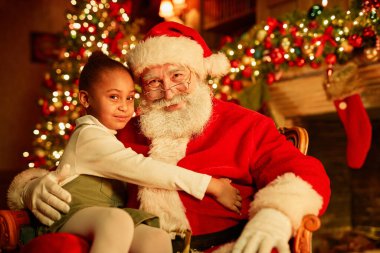 Geleneksel Noel Baba 'nın gülümseyen portresi ve kucağında Noel ağacının yanında oturan tatlı küçük kız.