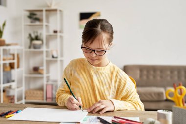 Portrait of teenage girl with Down syndrome drawing pictures at table in cozy room and smiling, copy space clipart