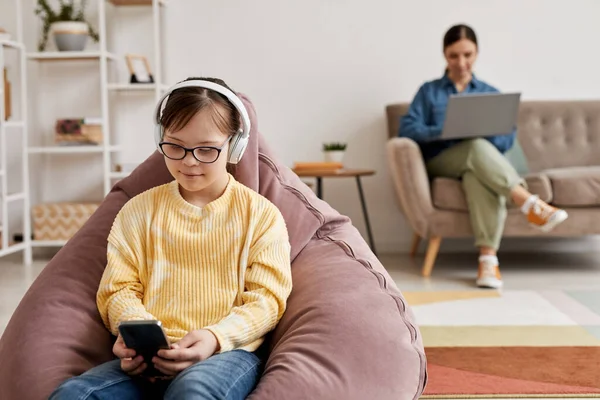 Portrait of teen girl with Down syndrome playing mobile games at home with parent supervision, copy space