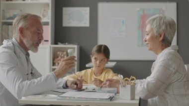 Orta yaşlı beyaz önlük giyen, beyaz çocuk doktoru, Asyalı küçük kızlara ilaç veren, geniş doktor ofisinde birlikte oturan orta yaşlı beyaz çocuk doktoru.