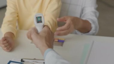 Klinikteki randevusu sırasında küçük Asyalı kızı ve annesini kontrol ederken kızıl ötesi termometre kullanan tanınmayan bir erkek doktorun omzundan.