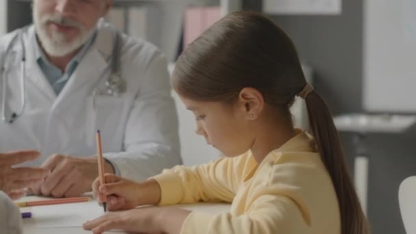 黄色いスウェットシャツの小さなアジアの女の子のチェストアップは 任命中に彼女の母親に話している間 医師のオフィスの机に座っている紙のシートに描きます — ストック動画