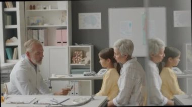 Klinikteki randevusu sırasında annesiyle birlikte küçük bir kızı kontrol ederken, beyaz laboratuvar önlüğü giyen, temassız kızılötesi termometre kullanan beyaz erkek çocuk doktorunun orta boy fotoğrafı.