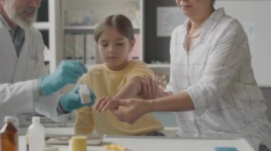 Orta yaşlı beyaz erkek doktor, laboratuvar önlüğü ve tıbbi eldiven giyiyor. Kolunda böcek ısırığı olan Asyalı küçük kıza ilk yardım yapıyor.
