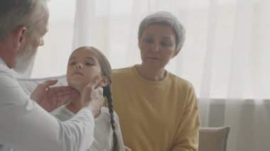 Orta boy beyaz erkek çocuk doktoru. Asyalı küçük kızın boynunu şişmiş bezler için inceliyor. Gündüz vakti annesiyle birlikte kanepede oturuyor.