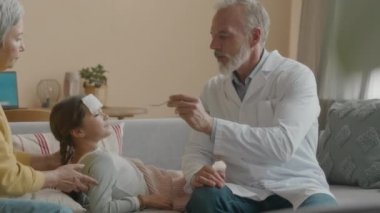 Beyaz laboratuvar önlüklü, beyaz erkek doktorun, büyükannesinin kucağında kanepede, alnında bez bezle küçük bir kıza ilaç şurubuyla kaşık verdiği orta boy bir fotoğraf.