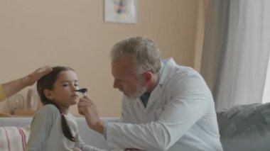 Orta yaşlı beyaz erkek doktor, ev ziyareti sırasında Asyalı hasta kızın burnunu inceliyor. Muayeneden sonra büyükannenin kucağına uzanan bir kız.