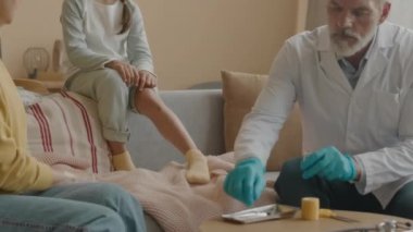 Orta boy erkek doktor iyota batırılmış pamuk çubuğu kullanırken küçük Asyalı bir kızın yaralı dizini tedavi ederken evde büyükannesiyle kanepede otururken.