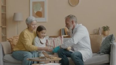 Orta yaşlı beyaz erkek doktor, laboratuvar önlüklü. Asyalı küçük bir kızın bacağını sargılıyor. Oturma odasında büyükannesiyle birlikte oturuyor.