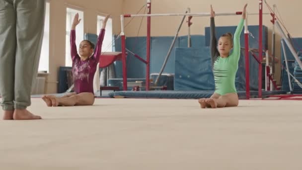 Kaksi Monietnistä Pikkutyttöä Trikooissa Tekemässä Venyttelyä Synkronoidusti Harjoitellessaan Rytmistä Voimistelua — kuvapankkivideo