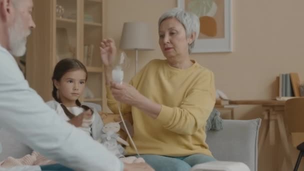 医師の家の訪問中に吸入器を使用する喘息アジアの少女のミディアムショット明るいリビングルームのソファに母親と座って — ストック動画