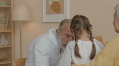 Asyalı küçük bir kızın kalbini dinlerken steteskop kullanan beyaz, olgun bir erkek çocuk doktoru.