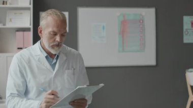 Beyaz laboratuvar önlüklü beyaz erkek doktorun orta boy portresi. Tıbbi dokümanlara not alıyor ve kameraya gülümsüyor. Modern doktorların ofisinde poz veriyor.