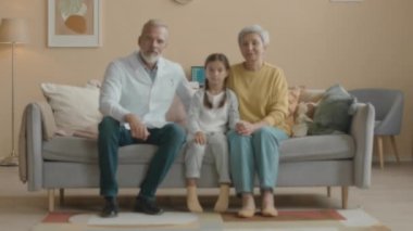 Beyaz laboratuvar önlüklü beyaz erkek çocuk doktorunun portresi. Küçük Asyalı kız ve annesi kanepede, rahat oturma odasında kameraya gülümsüyor.