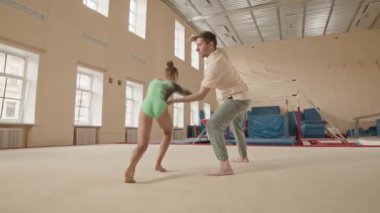 Geniş bir sınıfta antrenman yaparken jimnastik hocasıyla ön perende antrenmanı yapan yeşil taytlı, beyaz bir kız.