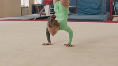 Konsantre olmuş, beyaz giysili küçük kız geniş bir sınıfta jimnastik yaparken sanatsal elementlerin zarif zemin rutinini yapıyor.