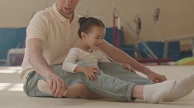 Profesyonel Kafkasyalı erkek jimnastik eğitmeni. Jimnastik tesisleriyle donatılmış geniş bir stüdyoda, kapalı alanda bacak esneten 5 yaşındaki Asyalı kıza yardım ediyor.