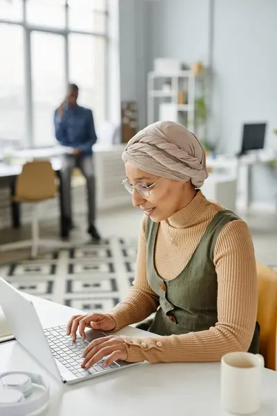 Vertical portrait of young Middle Eastern businesswoman wearing headwrap in office and typing at laptop