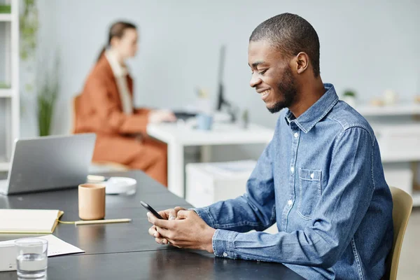 Minimal side view portrait of smiling black man using smartphone in office setting at break and chatting online