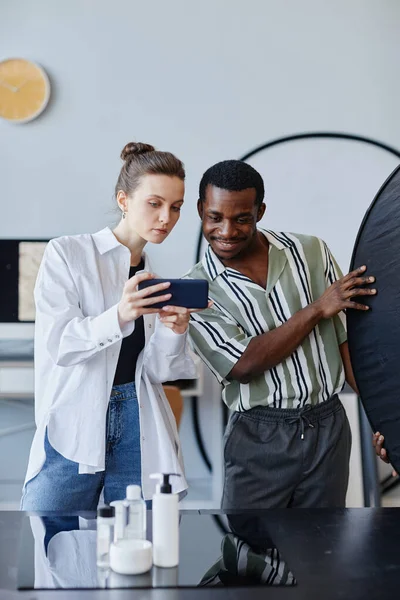 Vertical portrait of two young photographers working on product photography in studio and using mobile phone for social media
