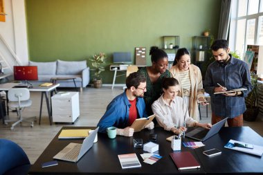 Genç çok kültürlü çalışanlar iş yerlerinin önünde toplanmış online videoları tartışıyor ve ofiste notlar alıyorlar.
