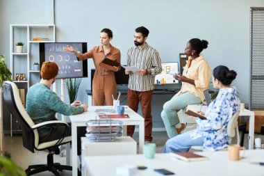 İki genç, kendine güvenen yönetici pazar oranı değişiklikleri hakkında rapor verirken, kadın ekonomist ekrandaki grafik verilere işaret ediyor.
