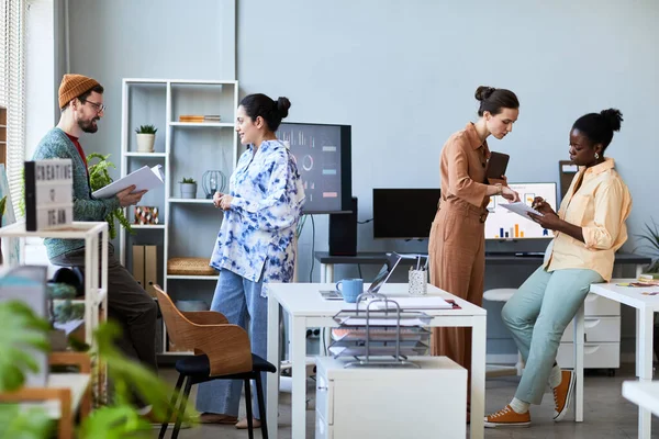 Two small group of young successful coworkers in casualwear discussing their ideas and notes while preparing for seminar in office