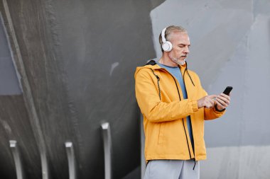 Dış mekânda kulaklık takan yakışıklı bir adamın yan görünüş portresi ve beton arka planda akıllı telefon ile müzik kurma, mekanı kopyalama