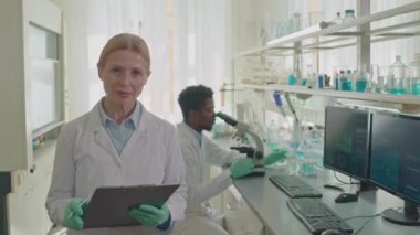 Beyaz kadın laboratuvar bilim adamının orta boy portresi. İş yerinde elinde panoyla kameraya poz veriyor. Siyah erkek araştırmacı cam şişede sıvı maddeyi inceliyor ve arka planda mikroskop kullanıyor.