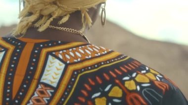 Sarı başörtüsü takan gülümseyen Afrikalı genç kadının yavaş yavaş yakın plan portresi, geleneksel parlak kıyafetler ve kamera için poz veren çöl manzarası bulanık arka plan