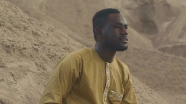 Orta karar, sarı tişörtlü Afro-Amerikan bir adam boş çölde kumların üzerinde otururken ileriye bakıyor.