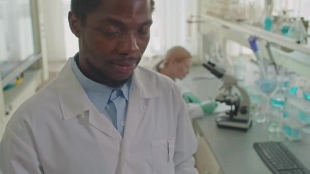 白人女性の同僚との研究室で仕事の日にカメラを見ている若い黒人男性研究室の科学者の肖像画をチェストアップ — ストック動画