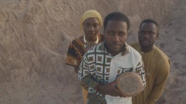 Üç kişilik Afro-Amerikan kabilesinin geleneksel süslenmiş giysileri içinde gün batımında kum tepeciğinde yan yana duran kameraya bakarken yüksek açılı portresi. Ortadaki adam elinde djembe tutuyor.