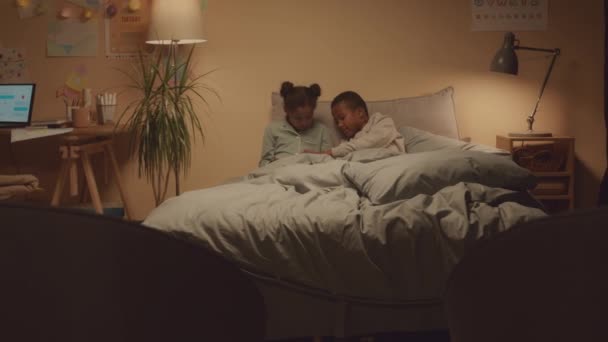 美国电话电报公司拍摄的非洲裔美国人兄妹穿着可爱的睡衣深夜坐在舒适的卧室里玩数字平板电脑游戏的照片 — 图库视频影像