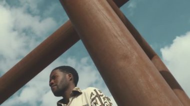 Geleneksel süslü gömlekler giyen düşünceli genç Afrikalı Amerikalı adam güneşli bir günde açık mavi gökyüzünün altında dikilirken etrafına bakıyor.