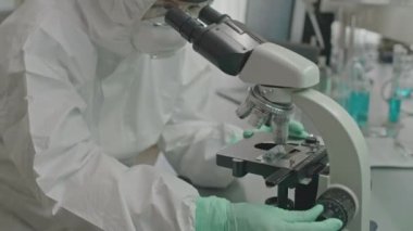 Modern laboratuarda mikroskop üzerinde çalışan solunum maskeli, güvenlik gözlüklü ve koruyucu örtülü kadın bilim adamı.