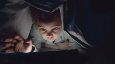 Afrika kökenli Amerikalı ergen kızın gece yatağında yumuşak bir battaniyenin altında yatarken el feneriyle dijital tablet kullanması.